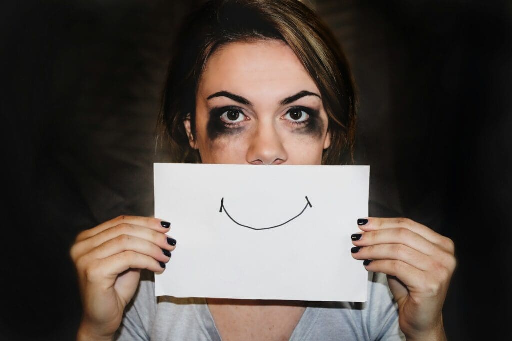 a woman with massive holiday stress, who has been crying (her eye make up is smeared) hides her worried face behind a paper with a smile drawn on it 