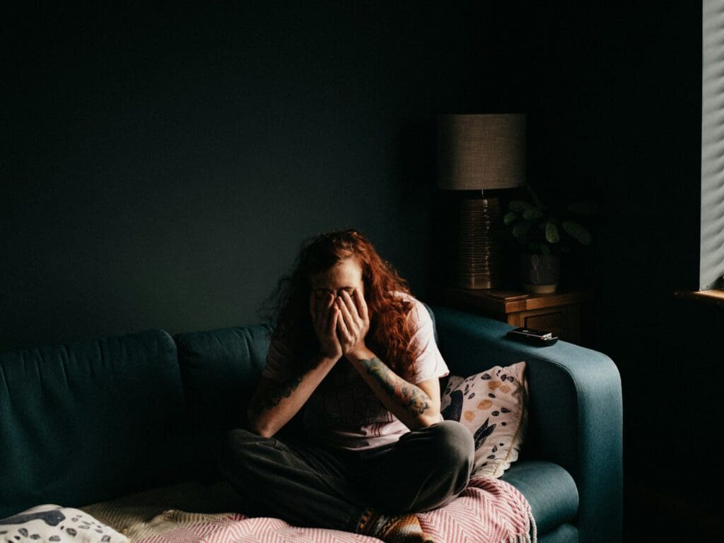 woman in black and white floral shirt sitting on blue couch in a darkened room. She covers her face in her hands. She is all alone