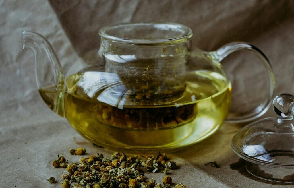 clear glass tea pot filled with hot water and dried chamomile flowers on a table top covered with more flowers