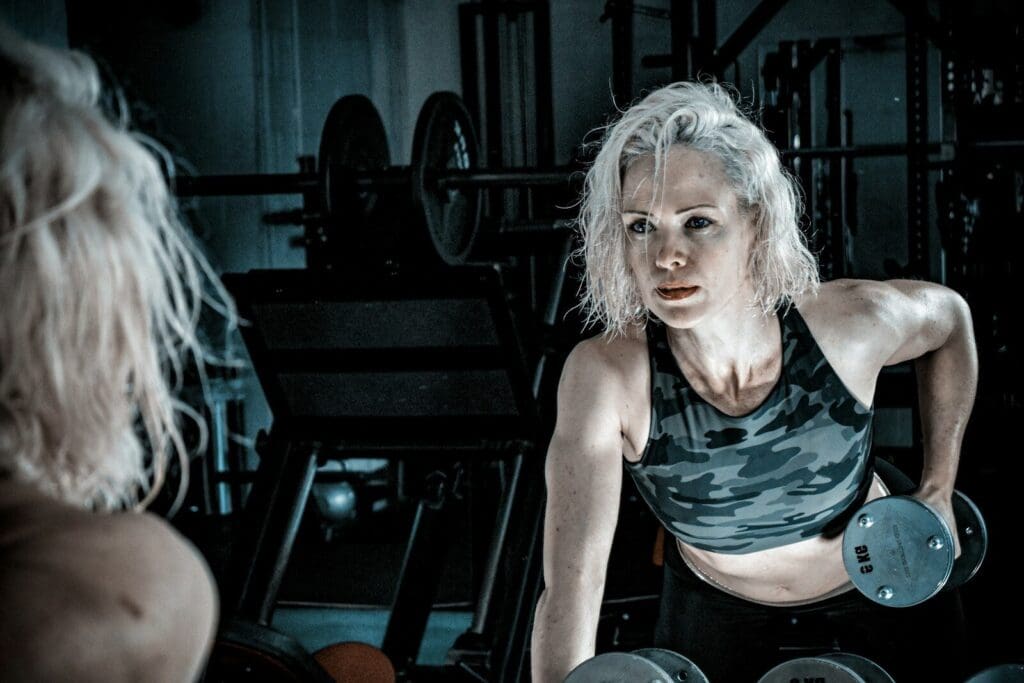 Older woman in black tank top lifting dumbbells as she checks form in the mirror. Strength traning over 50 is key to good health.