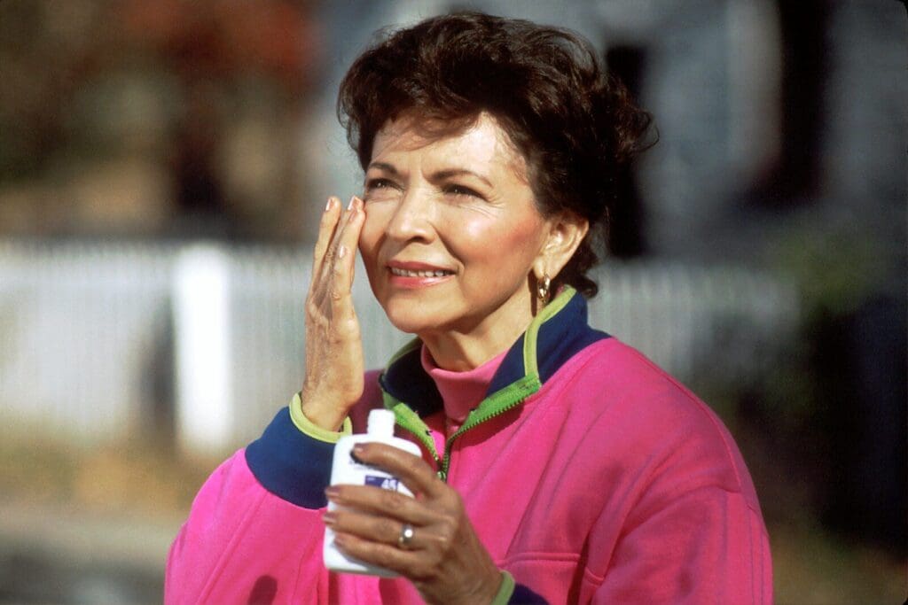 smiling woman in pink and blue shirt applies sunscreen to her face