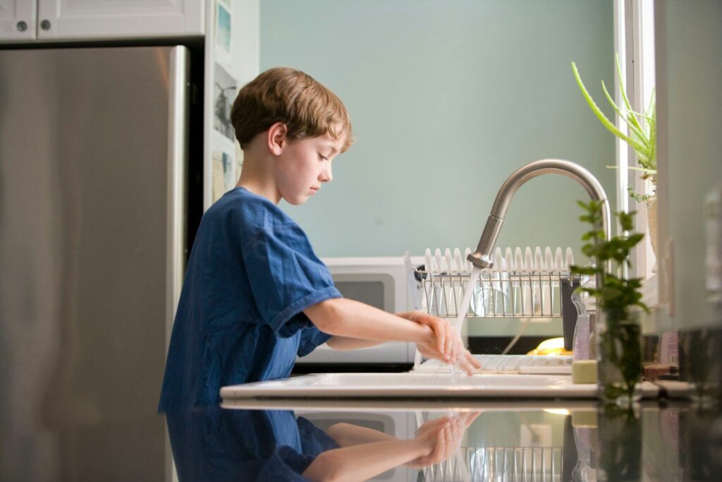 Young blonde boy in blue crew neck t-shirt standing at a kitchen sink washing his hands. A great way to stay healthy this back to school season.