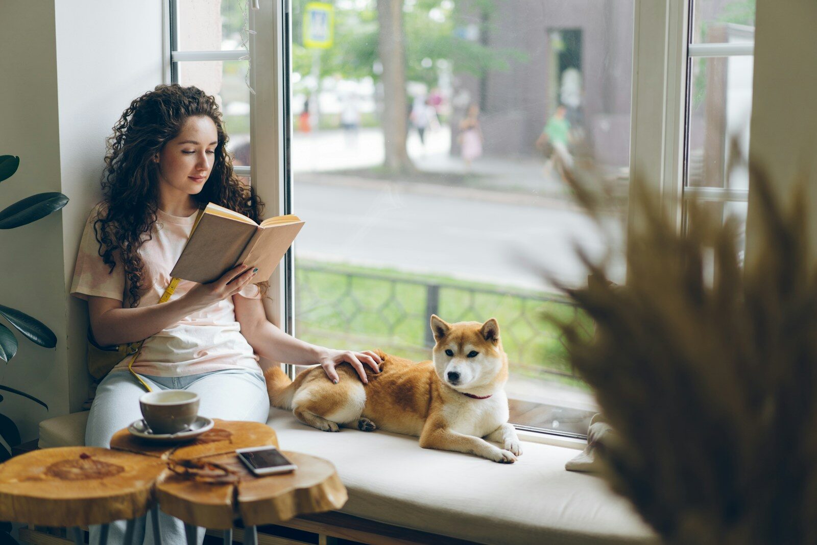 a white woman with dark hair sitting on a window sill reading a book next to a dog. Dogs are great stress relievers.